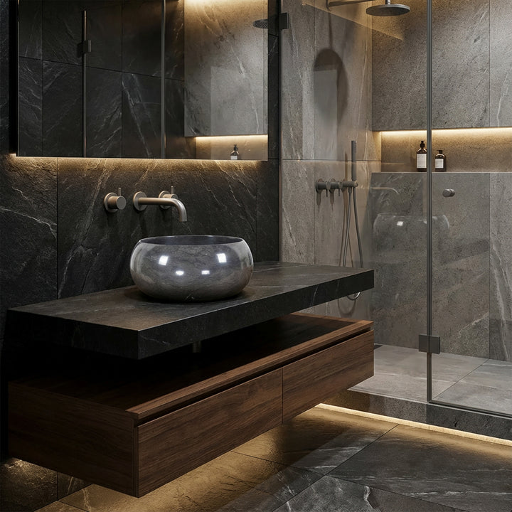 Lavabo sobre encimera redondo de piedra natural sobre un tocador oscuro en el moderno baño del spa, combinado con accesorios de pared, ducha de cristal e iluminación indirecta