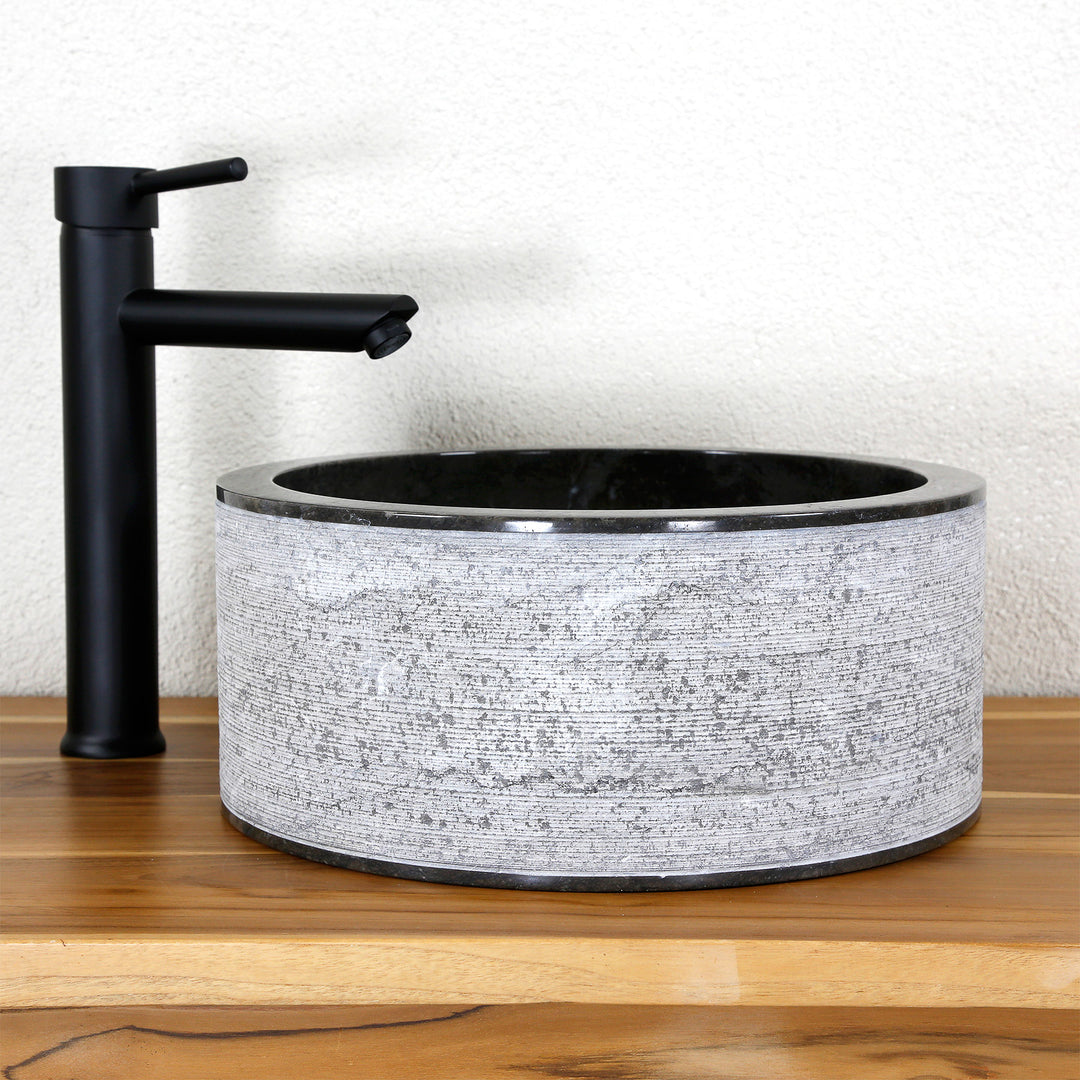 Lavabo sobre encimera redondo fabricado en piedra natural con una llamativa estructura de líneas, combinado con una grifería de pie sobre madera de color negro.