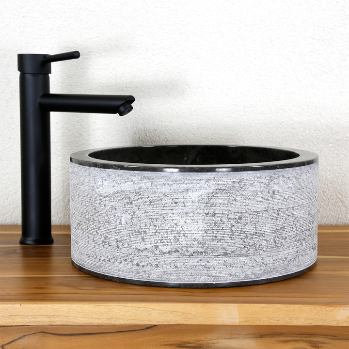 Lavabo sobre encimera redondo fabricado en piedra natural con una llamativa estructura de líneas, combinado con una grifería de pie sobre madera de color negro.