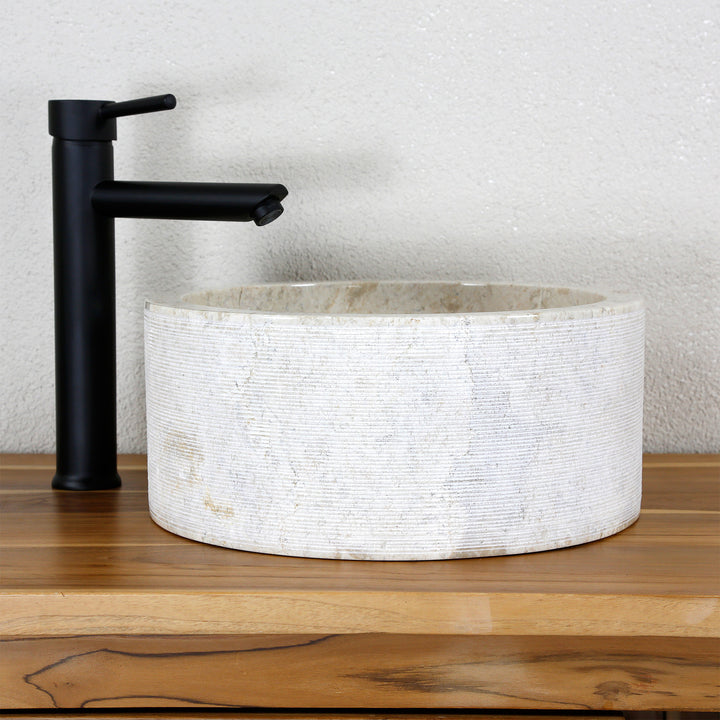 Lavabo sobre encimera redondo fabricado en piedra natural clara y estructura de líneas finas, combinado con grifería independiente de color negro.