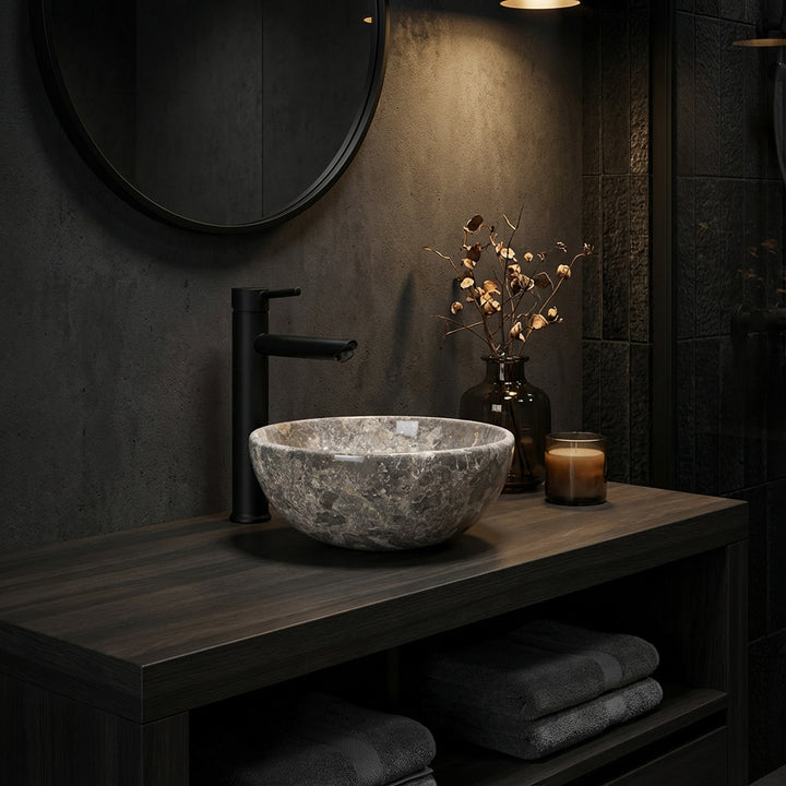 Lavabo sobre encimera redondo de piedra natural con vetas de color marrón grisáceo sobre una consola de madera oscura en el cuarto de baño con una iluminación agradable, combinado con grifería negra, espejo y accesorios sutiles