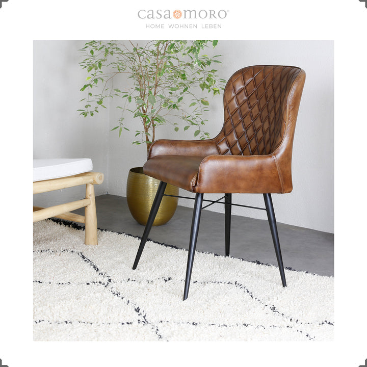 Antikbrauner Lederstuhl mit Rautensteppung auf hellem Teppich, daneben eine goldene Vase mit Grünpflanze – modernes Wohnambiente mit natürlicher Note.