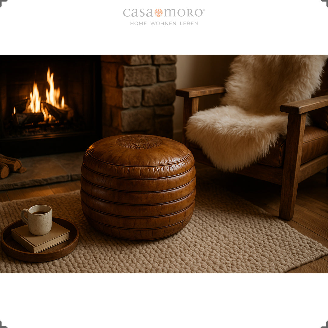 Gemütliche Wohnszene mit rundem Leder Pouf in warmem Braun vor dem Kamin, daneben ein Holzsessel mit Fellauflage und eine Tasse auf Tablett – stimmungsvolles Ambiente.