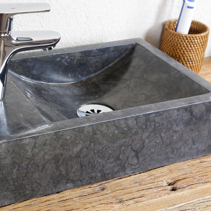 Lavabo sobre encimera angular de piedra natural con vetas oscuras en una vista detallada, con desagüe de acero inoxidable y grifería cromada sobre placa de madera