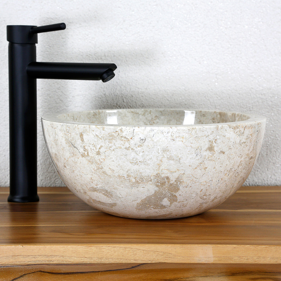 Lavabo de encimera redondo hecho de piedra natural clara sobre una tapa de madera con un accesorio negro en un sencillo ambiente de estudio