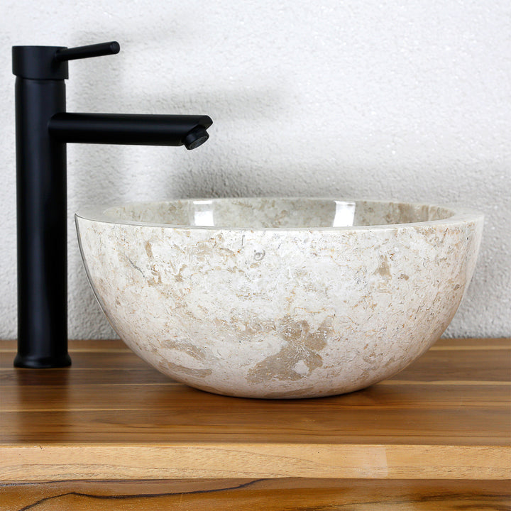 Lavabo de encimera redondo hecho de piedra natural clara sobre una tapa de madera con un accesorio negro en un sencillo ambiente de estudio