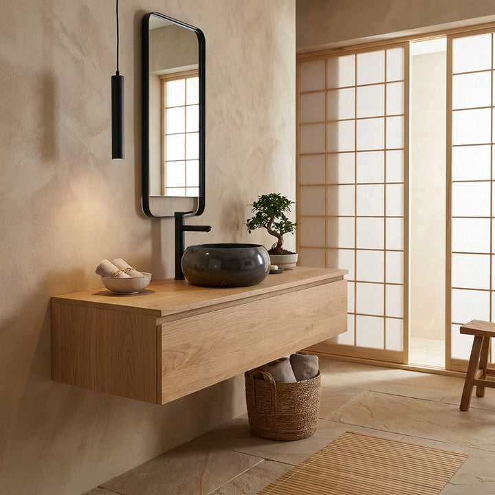 Lavabo sobre encimera de piedra redondo sobre tocador flotante de madera en un baño de inspiración japonesa, combinado con accesorios negros, espejo con marco de madera y suave luz natural