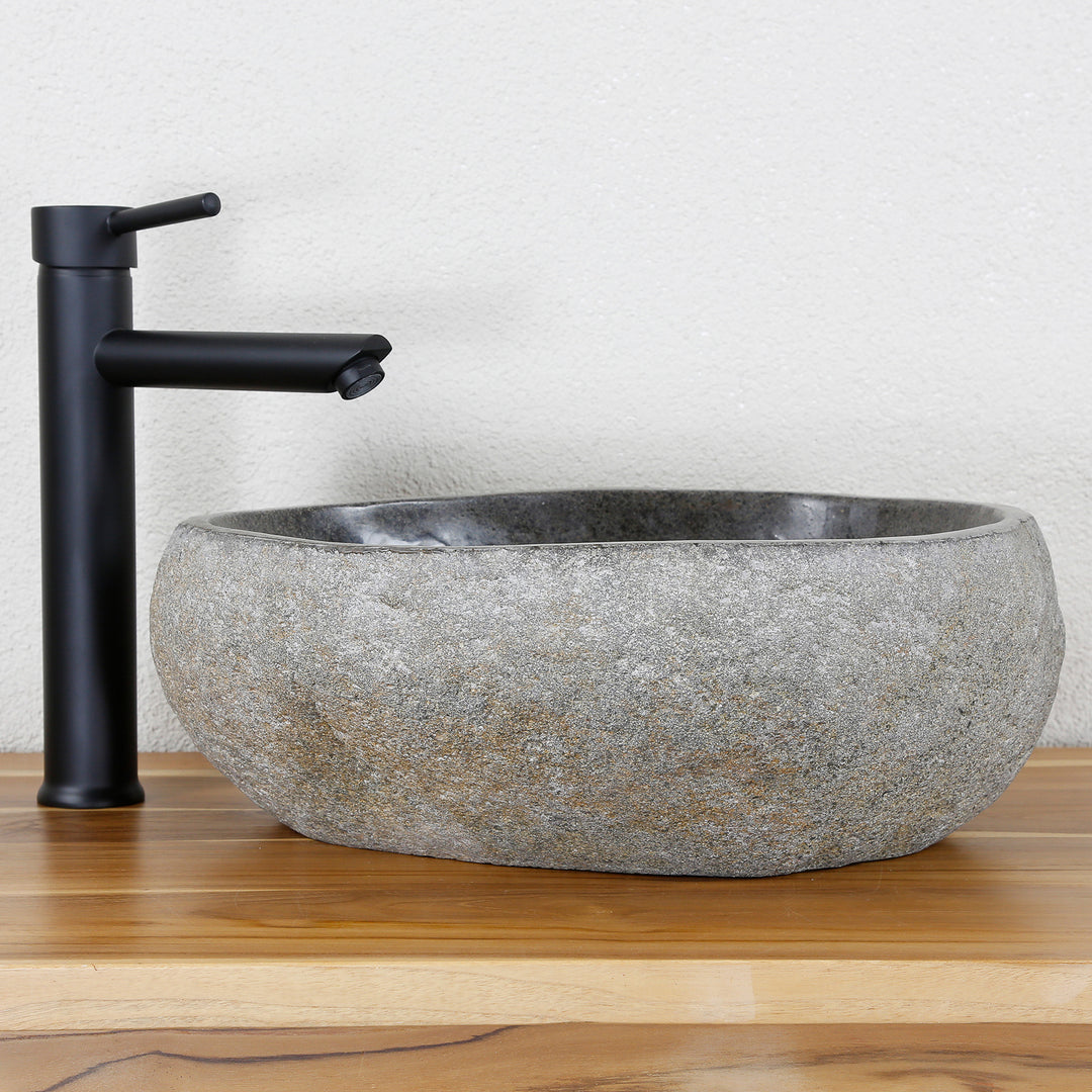 Lavabo Realizado en piedra natural gris de forma ovalada, combinado con herrajes negros sobre una cálida superficie de madera.