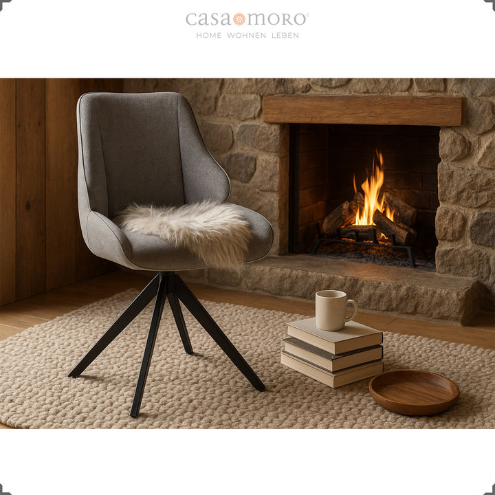 360° drehbarer Esszimmerstuhl in Grau vor Kamin mit Fellauflage, Bücherstapel und Tasse – gemütliche Wohnszene mit warmem Licht