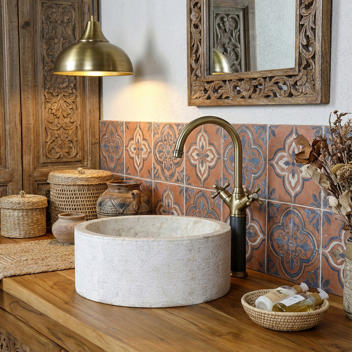 Lavabo redondo de piedra con un cálido aspecto de mármol sobre una consola de madera, complementado con herrajes de latón y azulejos orientales.