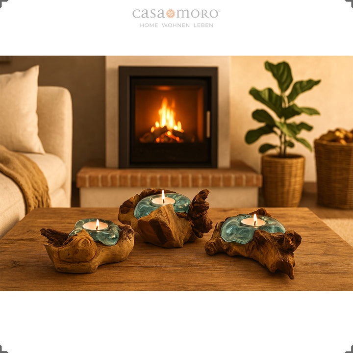 Drei Teelichthalter aus Glas und Wurzelholz auf einem Holztisch, ihr warmes Licht ergänzt die gemütliche Kaminatmosphäre im Wohnzimmer.