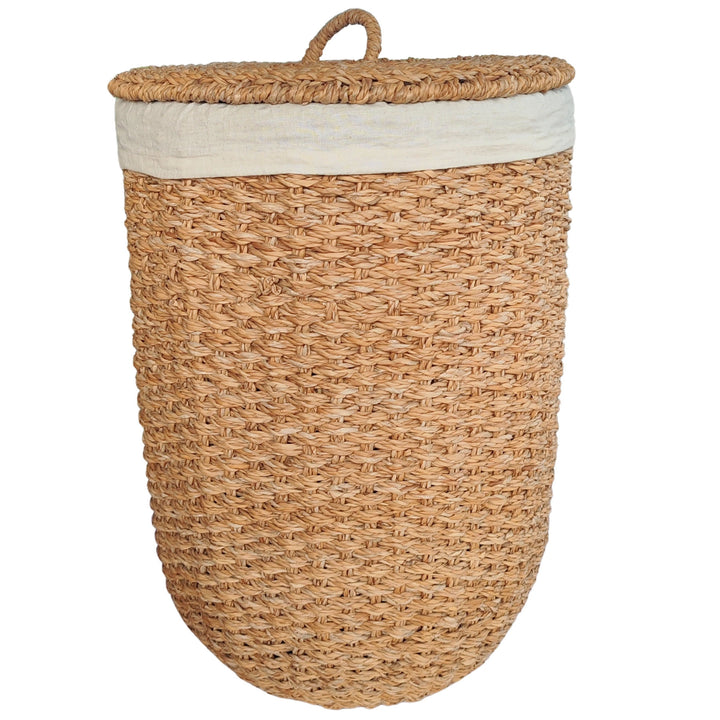 Cesto de la ropa abierto, fabricado en algas marinas de color natural, con un inserto de tela clara y una tapa apoyada lateralmente.