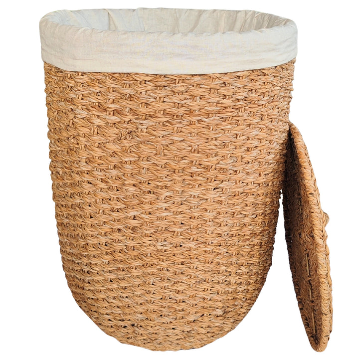 Cesto de la ropa abierto, fabricado en algas marinas de color natural, con un inserto de tela clara y una tapa apoyada lateralmente.