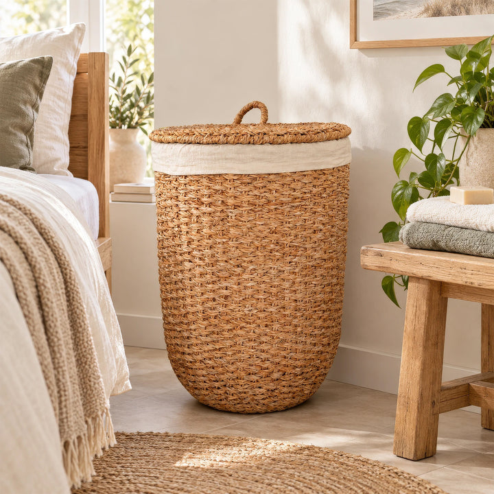 Cesto de la ropa con tapa, fabricado con algas marinas de color natural y con un inserto de tela clara, situado entre la cama y el banco de madera en el dormitorio.