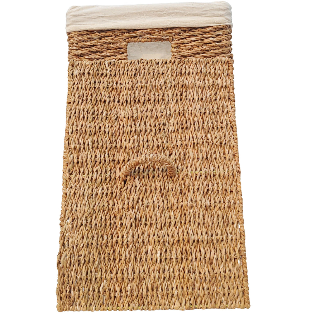 Cesto rectangular para la ropa, fabricado en algas marinas, con una capacidad de 90 litros, un forro de tela clara y una tapa con asa que se apoya en la parte delantera.
